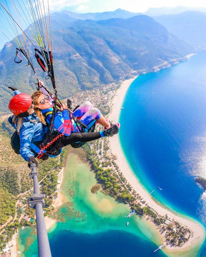 Two girls tandem paragliding over a turquoise blue lagoon and beach with mountains in the background