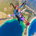 Two girls tandem paragliding over a turquoise blue lagoon and beach